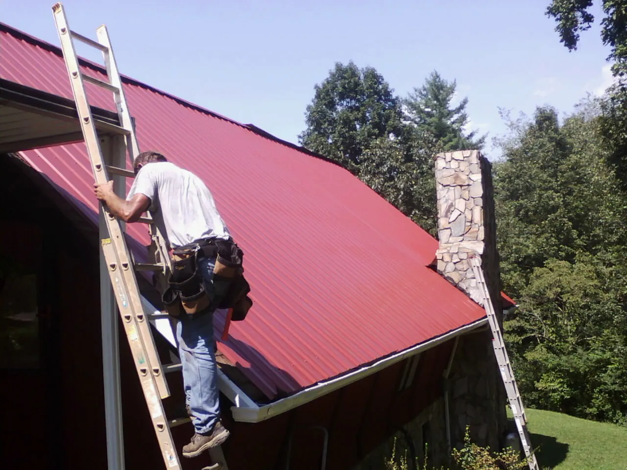 Roofer on a ladder beside a red metal roof and stone chimney — Soffit & Fascia Repair by Roofing Brothers in Havertown